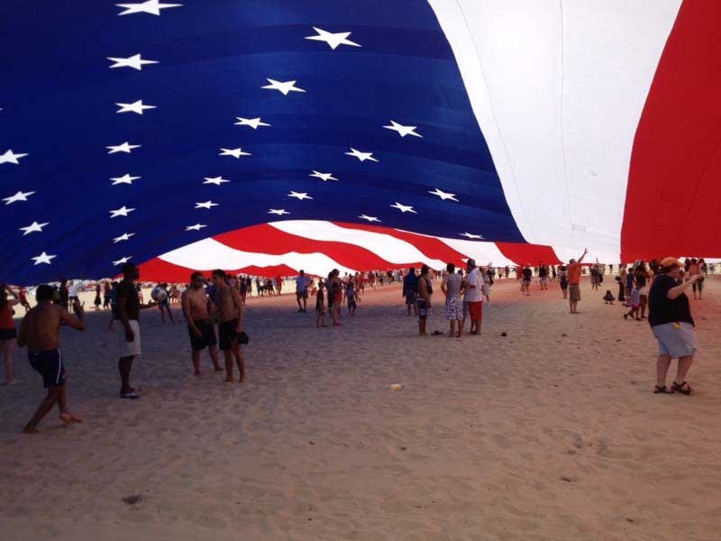 a large american flag on the beach with people walking around